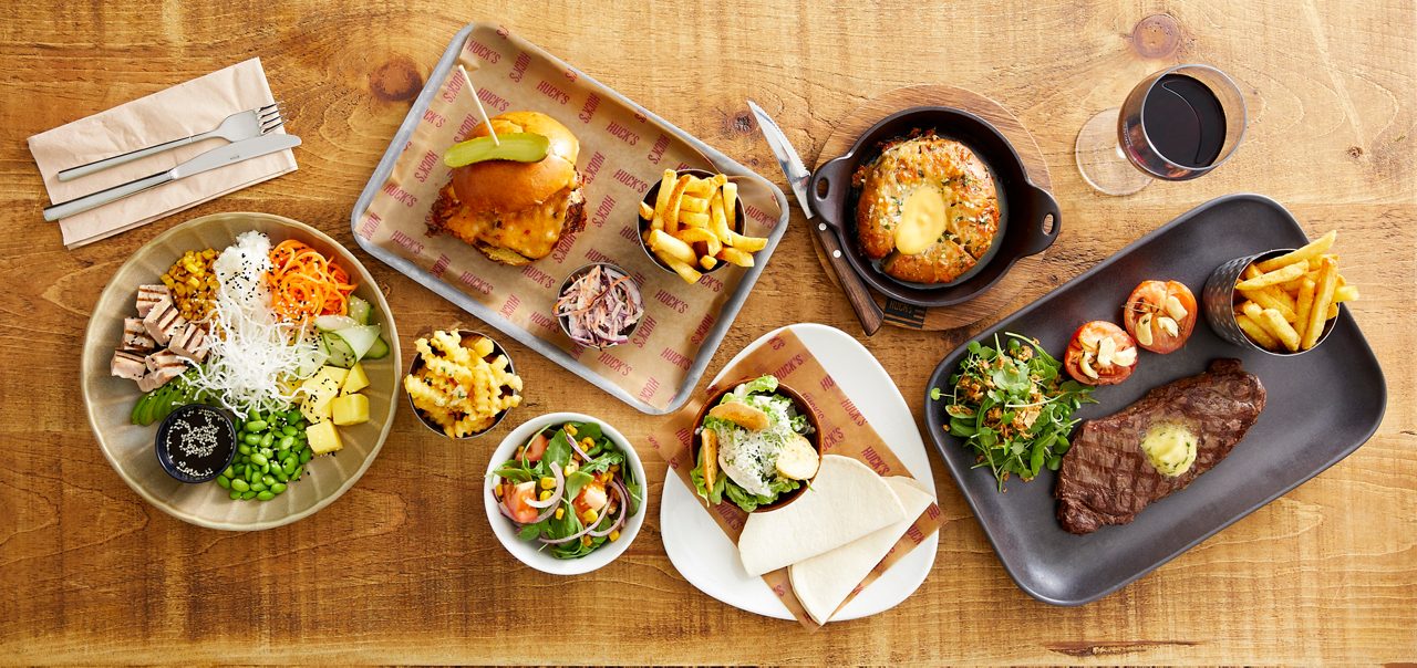 Assorted restaurant dishes sit arranged on a wooden table: steak with fries, burger with pickle, poke bowl, salads, mac and cheese, bread bowl dip, wraps, grilled tomatoes, glass of red wine. Text: repeated "HUCK'S" on paper liners.